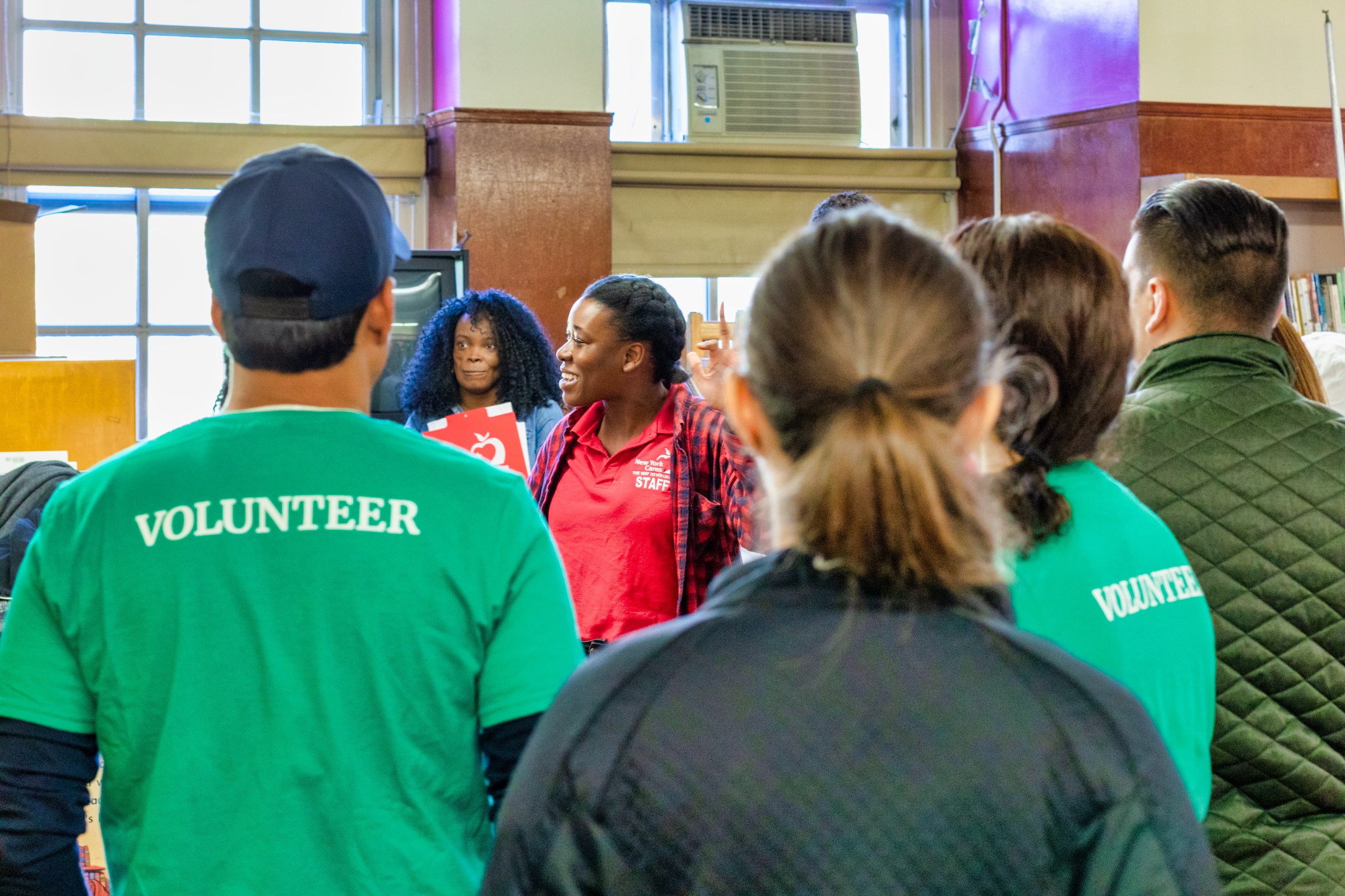 A group of 5 volunteers stand in a school gym with their backs to the camera. On one shirt text reads: "Volunteer"