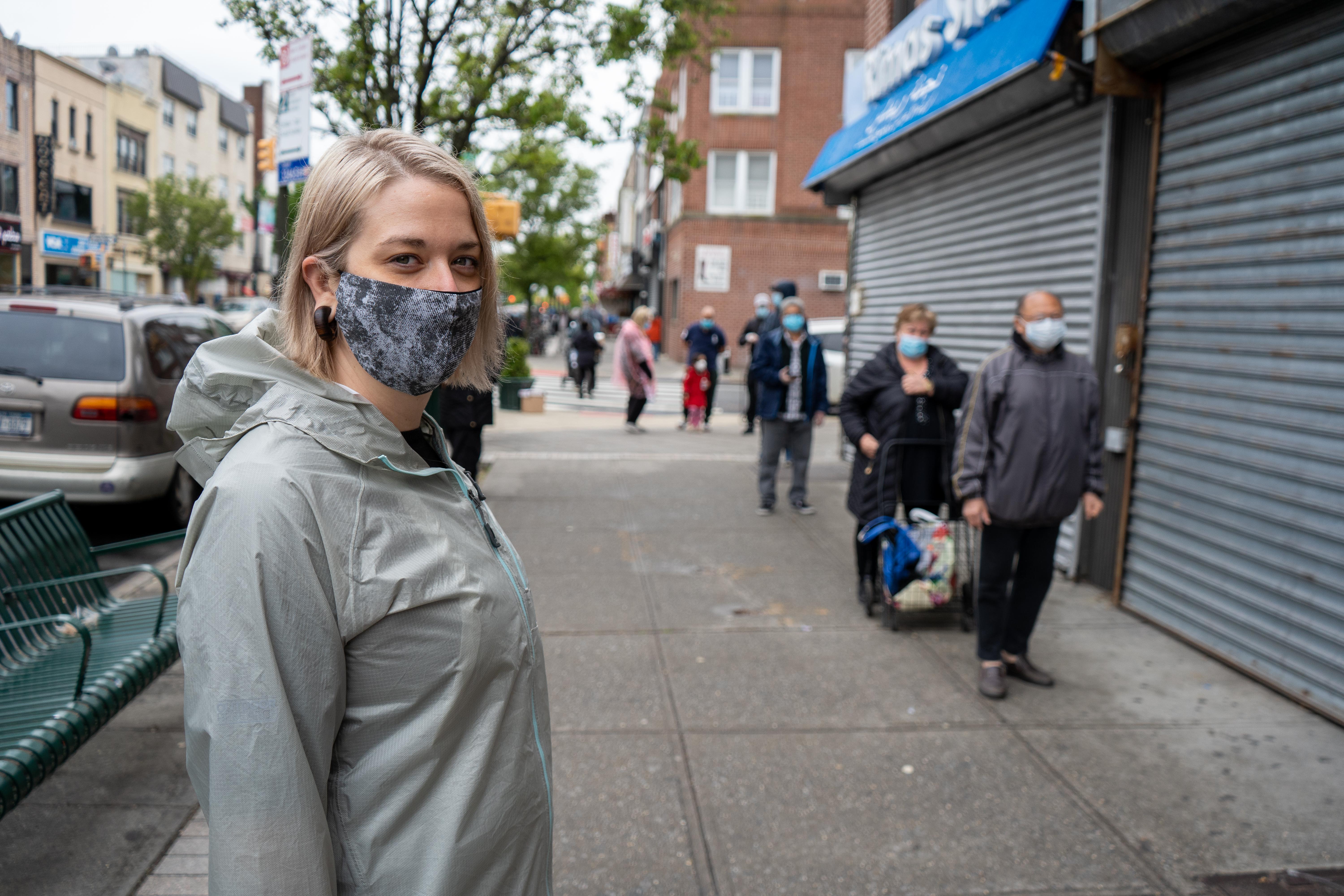A woman in a mask stands in the foreground on a city sidewalk and looks at the camera next to a line of others in masks