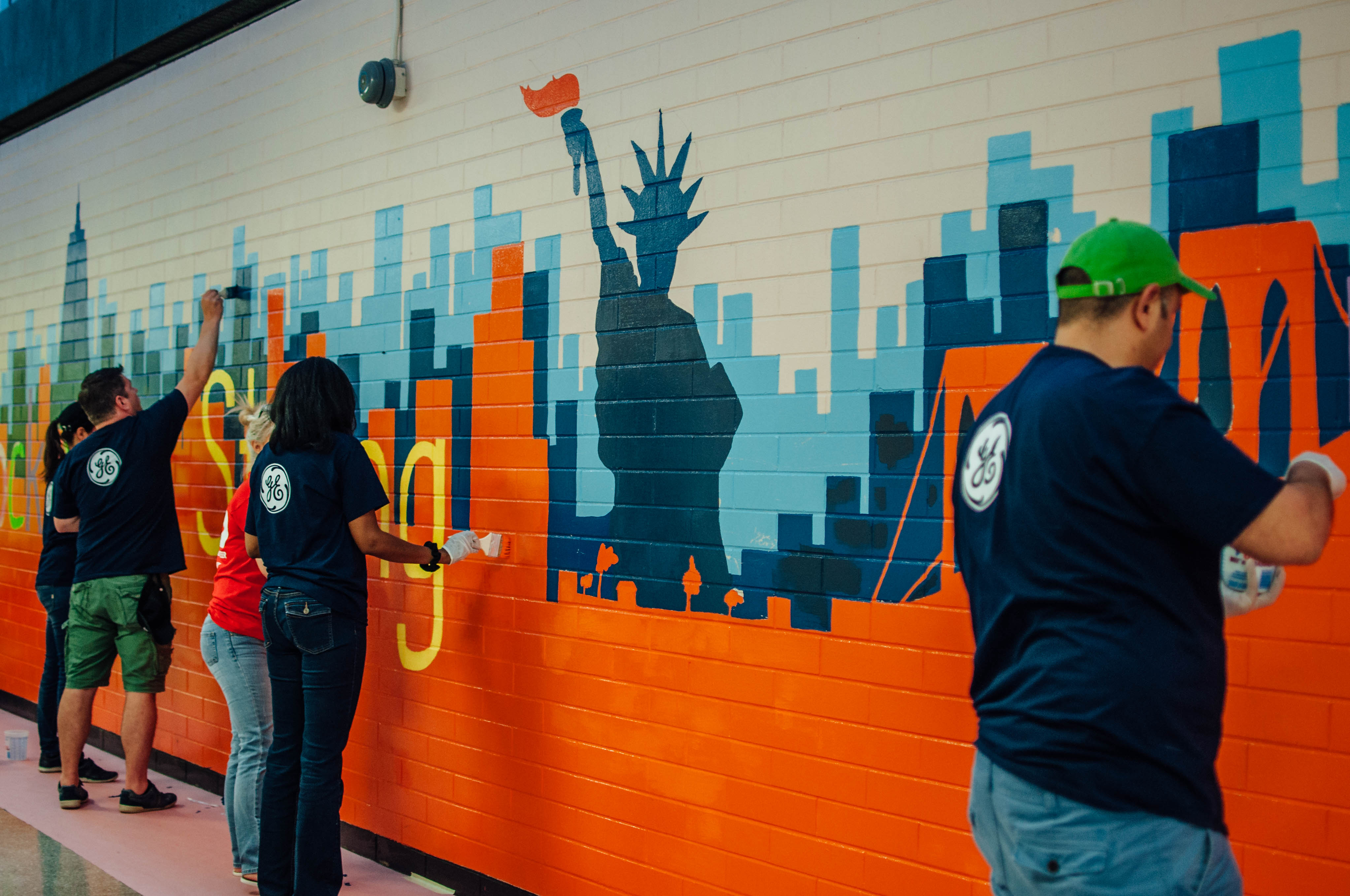 Four volunteers in matching blue shirts paint a mural on a school wall. The mural includes the Statue of Liberty.